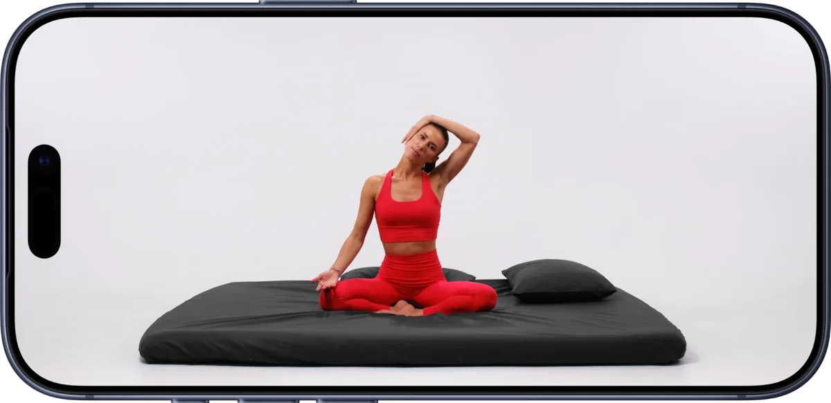 A phone turned sideways playing a bed session video: a woman sitting cross-legged on a mattress next to a pillow, stretching her neck to the side